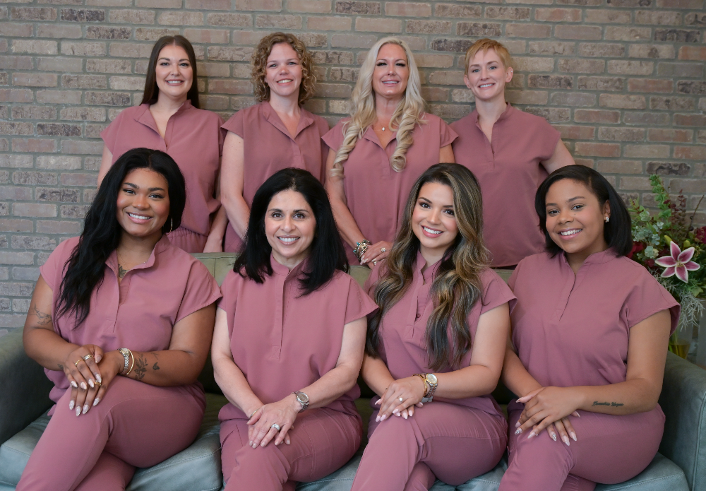 Group photo of Dr. Veena Bhat and her all-female dental team at North Fulton Smiles, all wearing matching mauve uniforms and smiling in front of a brick wall – North Fulton Smiles